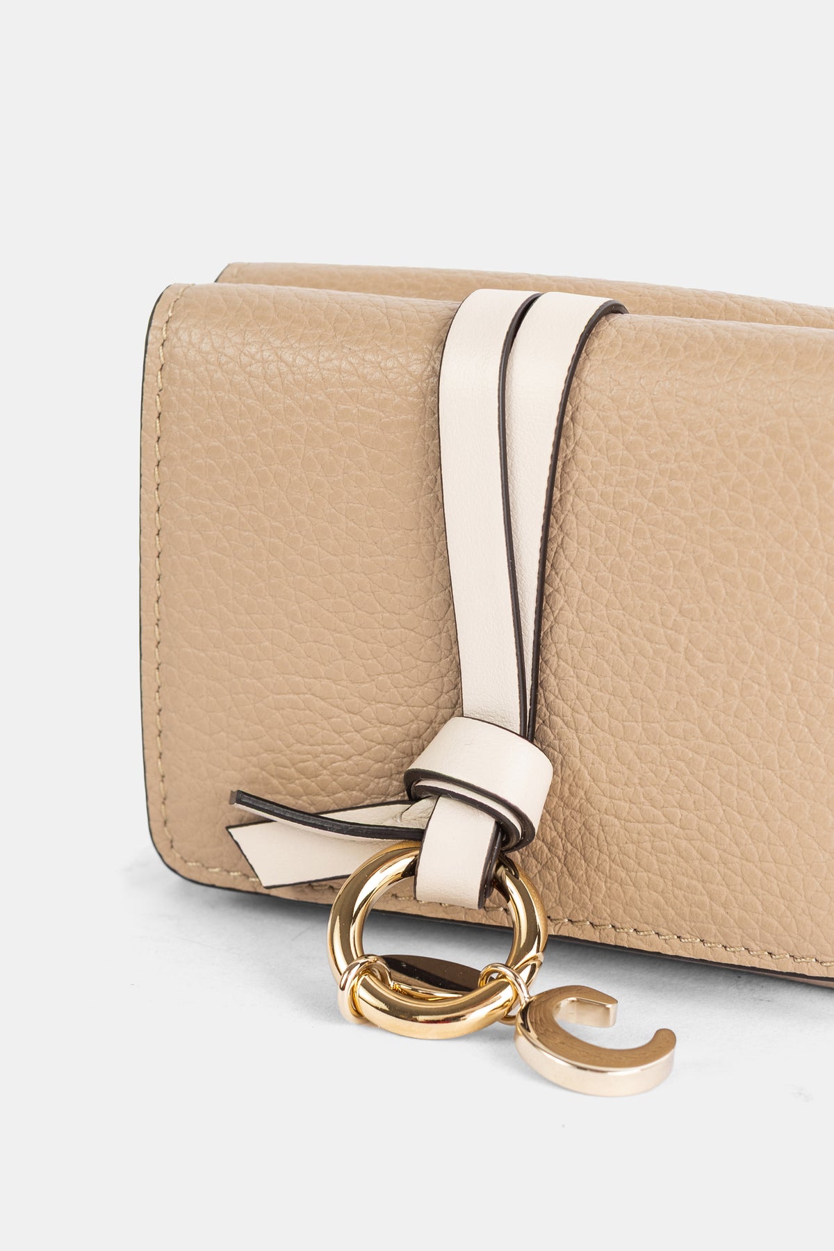 Pormenor da carteira Chloé Alphabet com berloque de logótipo dourado e tira de pele creme num fundo bege.