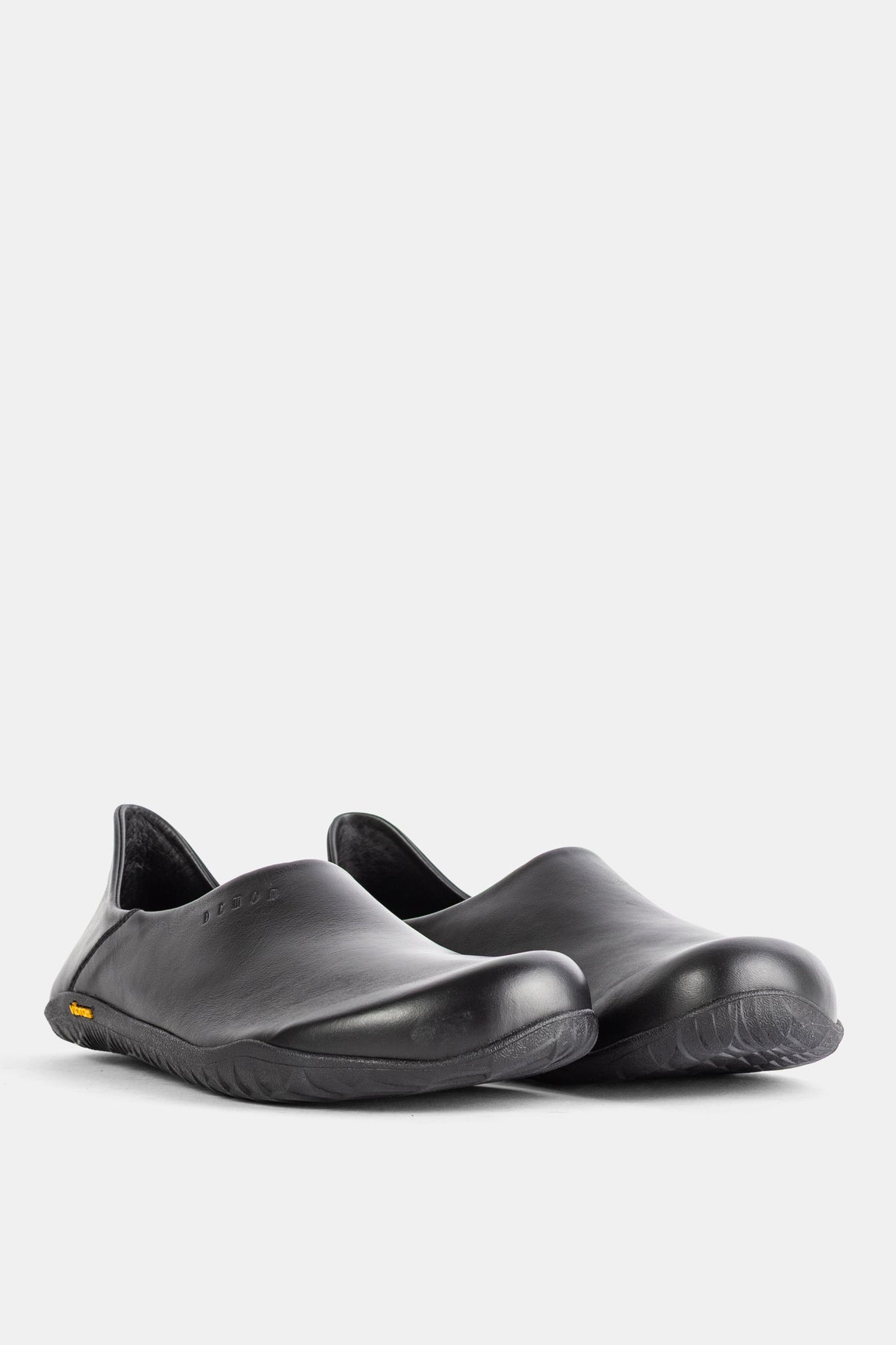 A pair of Demon Dalmara shiny black leather mules featuring an embossed logo and yellow Vibram sole details.