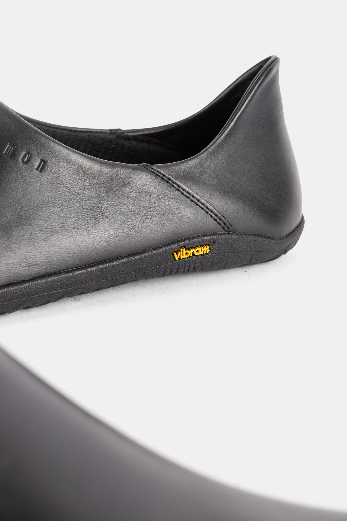 Rear heel view of the Demon Dalmara black leather mule with yellow Vibram logo on a slim minimalist sole.