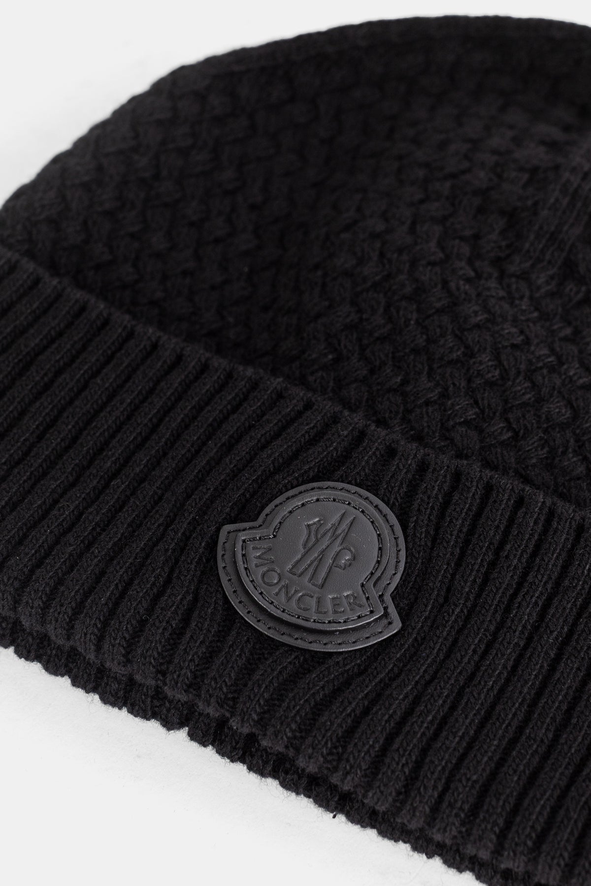 Detailed close-up of the rubber logo patch sewn onto the wool ribbed cuff of a black Moncler men's beanie.
