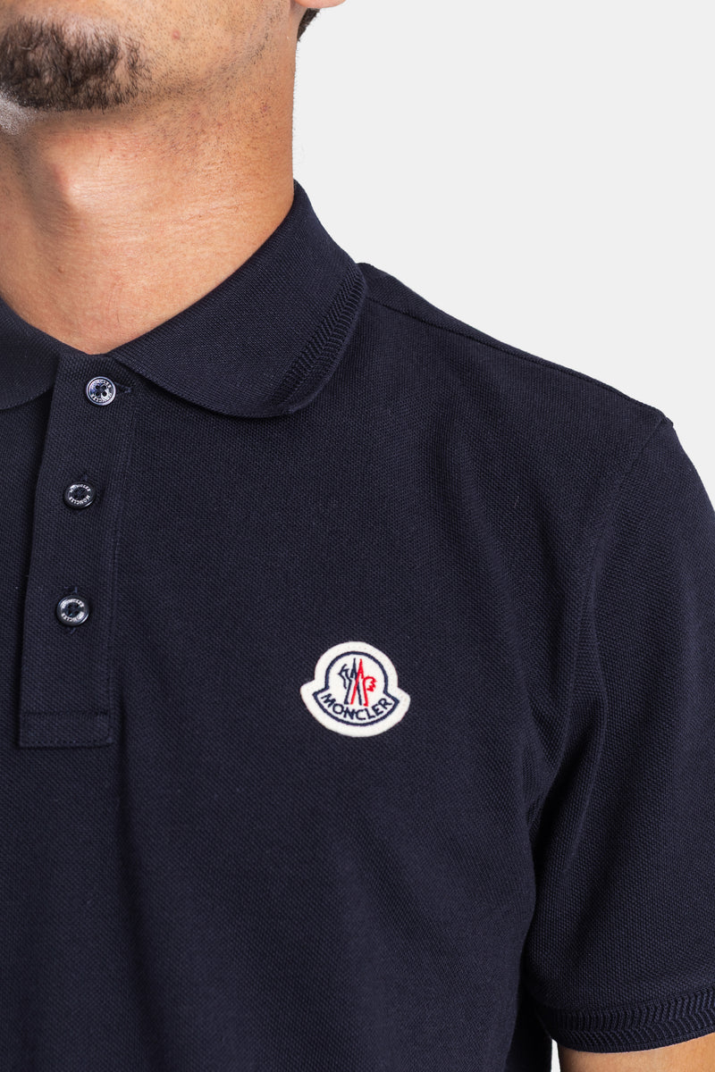 Close-up of Moncler men's navy cotton piqué polo featuring the round chest logo patch and matching buttons.