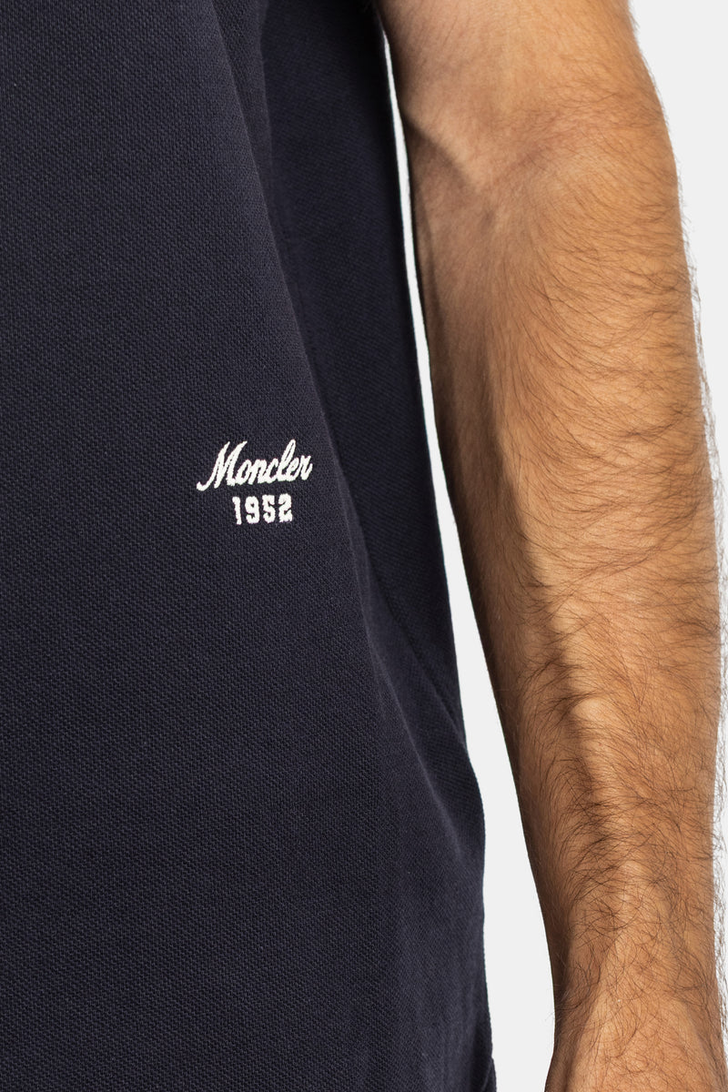 Close-up of Moncler navy blue polo's piqué cotton texture, white embroidered vintage script logo detail.