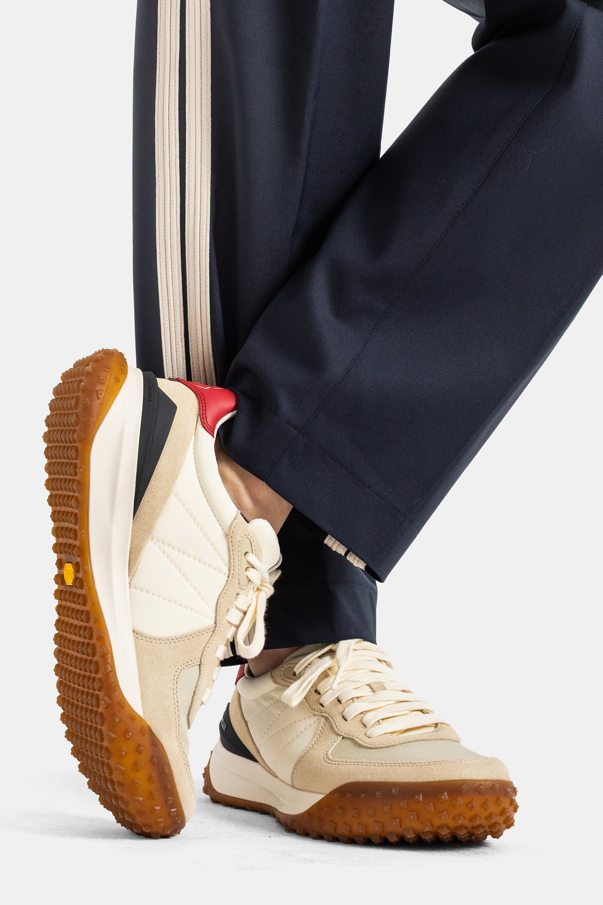 Moncler men's trousers close-up on navy leg hem with cream stripe, beige running sneakers, caramel rubber sole.
