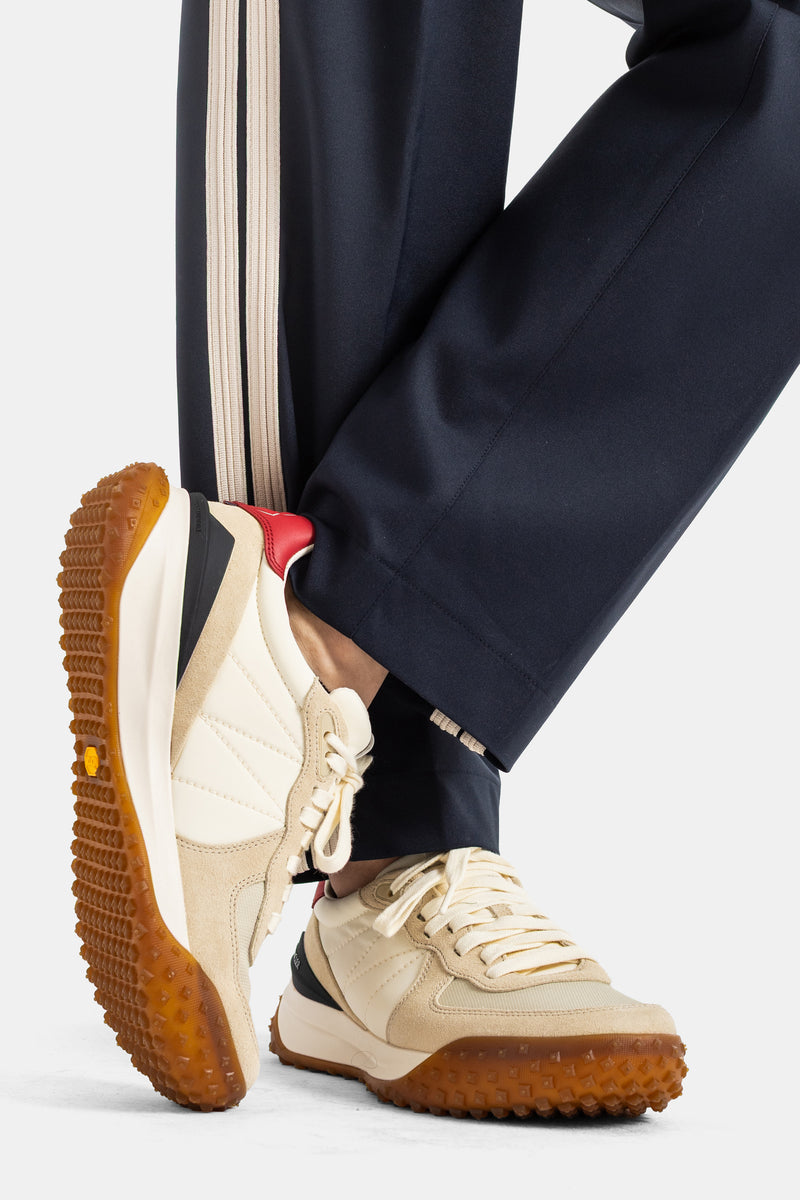 Moncler men's trousers close-up on navy leg hem with cream stripe, beige running sneakers, caramel rubber sole.