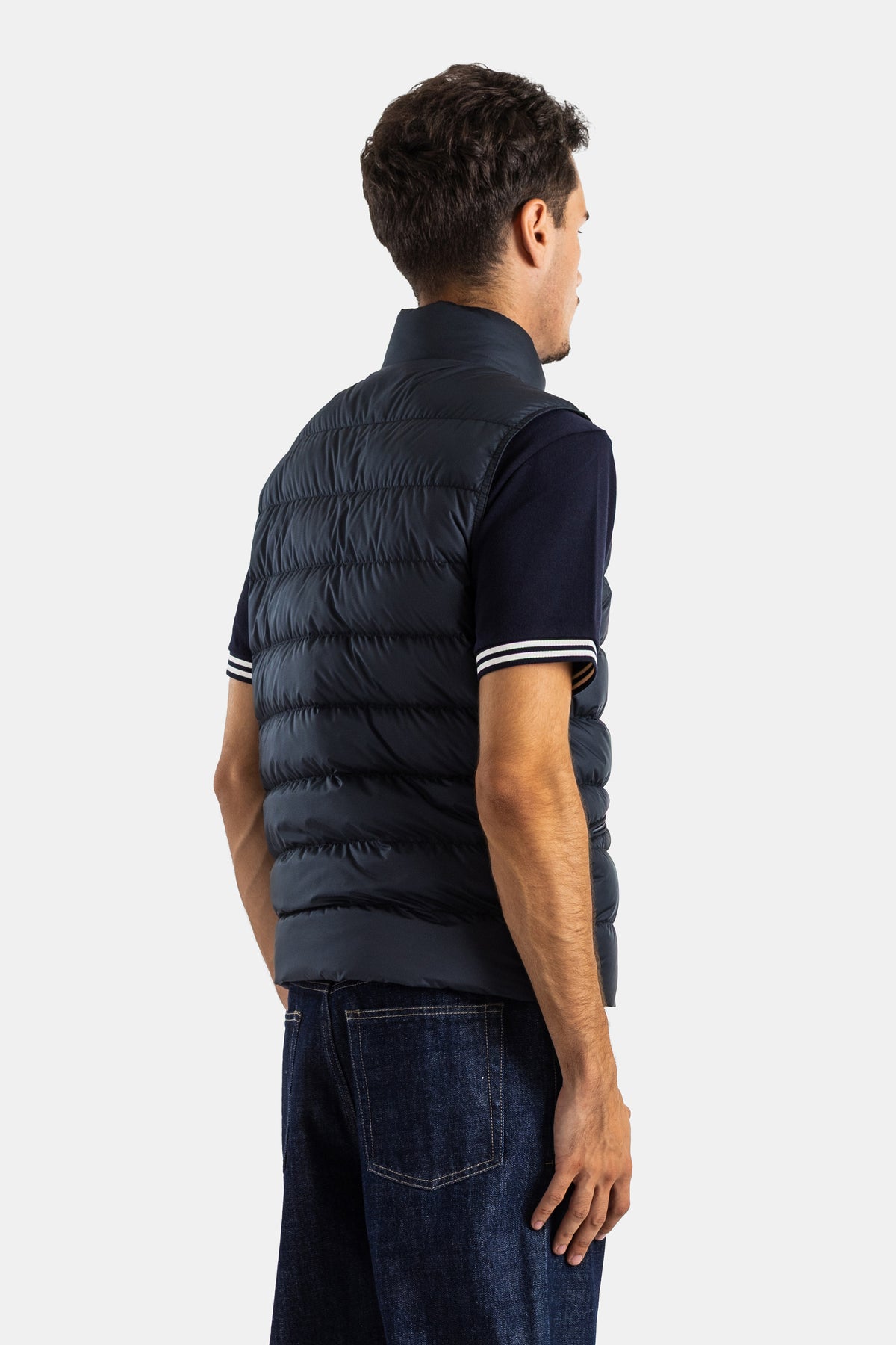 Back view of the Moncler Treompan men's navy blue sleeveless down jacket with horizontal padding and raw jeans.