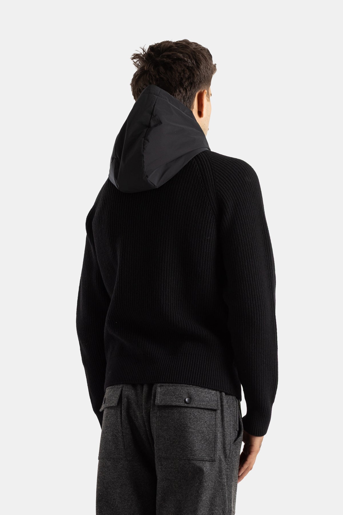 Back view of Moncler men's black ribbed knit cardigan, contrasting nylon hood, styled with grey wool pants.