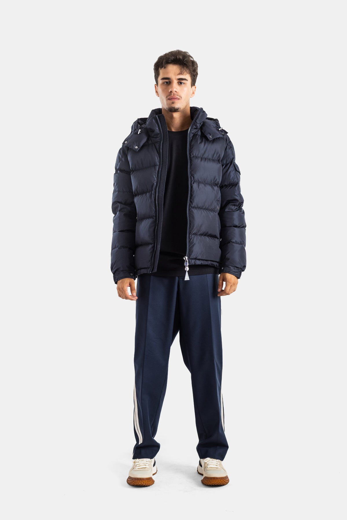 Moncler Maya men's total navy look with short matte down jacket worn open over black t-shirt and striped pants.