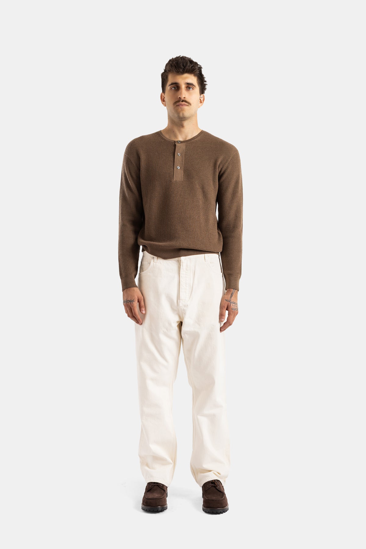 Vorderansicht der ecrufarbenen Studio Nicholson Jeans, getragen mit braunem Henley-Feinstrick und Wildlederschuhen.