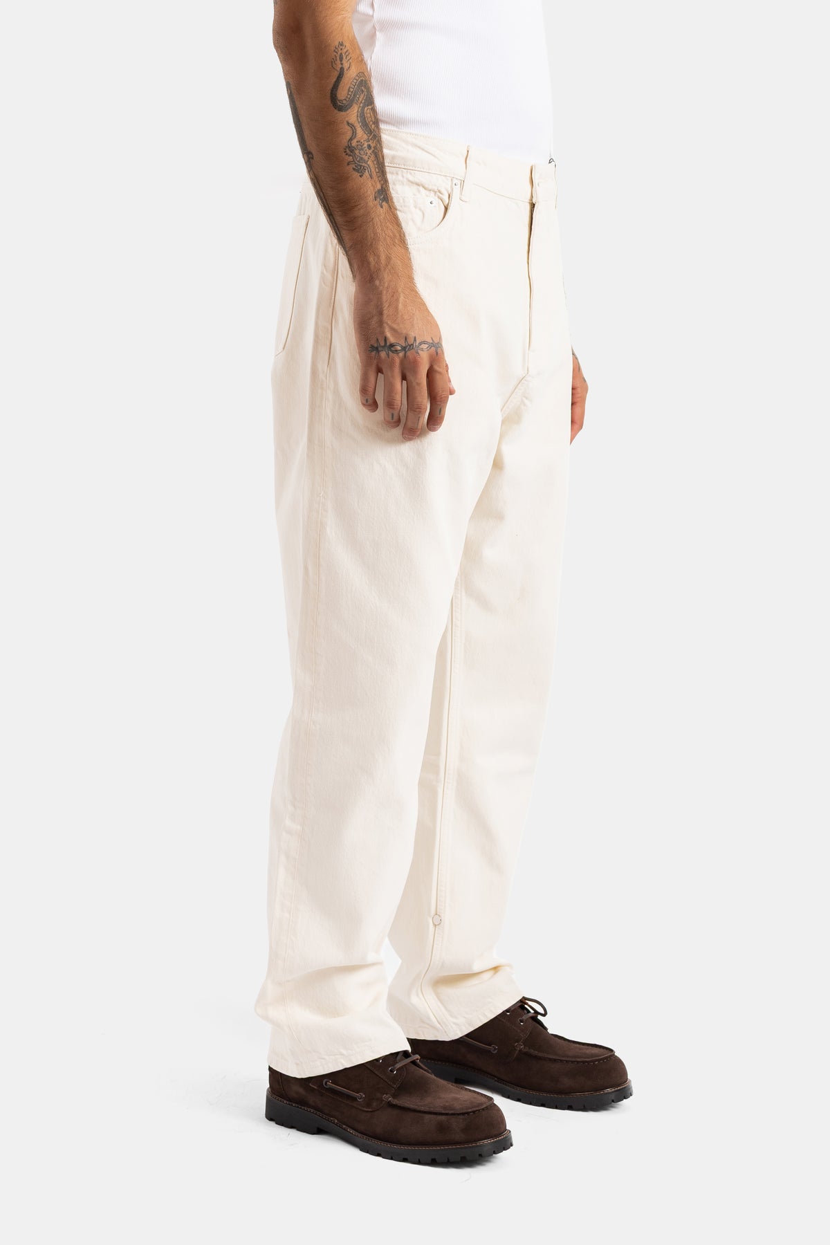 Seitliche Ansicht der hohen ecrufarbenen Studio Nicholson Jeans mit Bundfalten und braunen Wildleder-Mokassins.