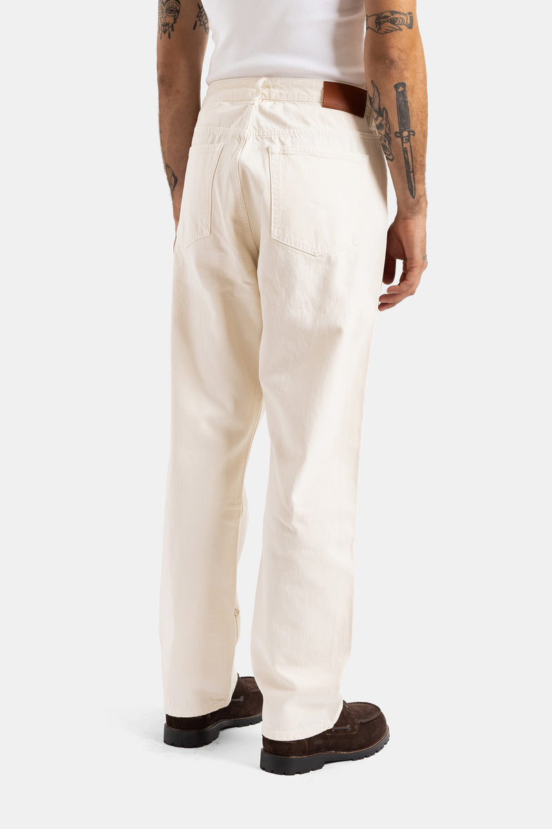 Rückansicht der ecrufarbenen Studio Nicholson Jeans mit Gesäßtaschen, cognacfarbenem Gürtel und tätowiertem Arm.