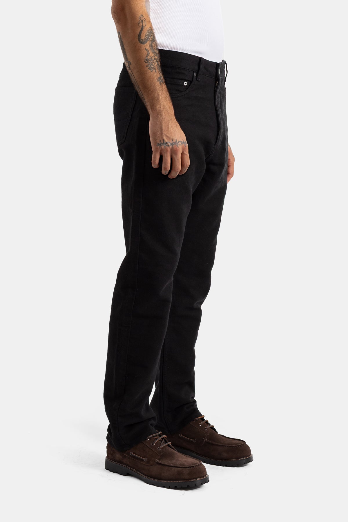 Studio Nicholson men's high-waisted black jeans in profile view with stylish brown suede boat shoes.