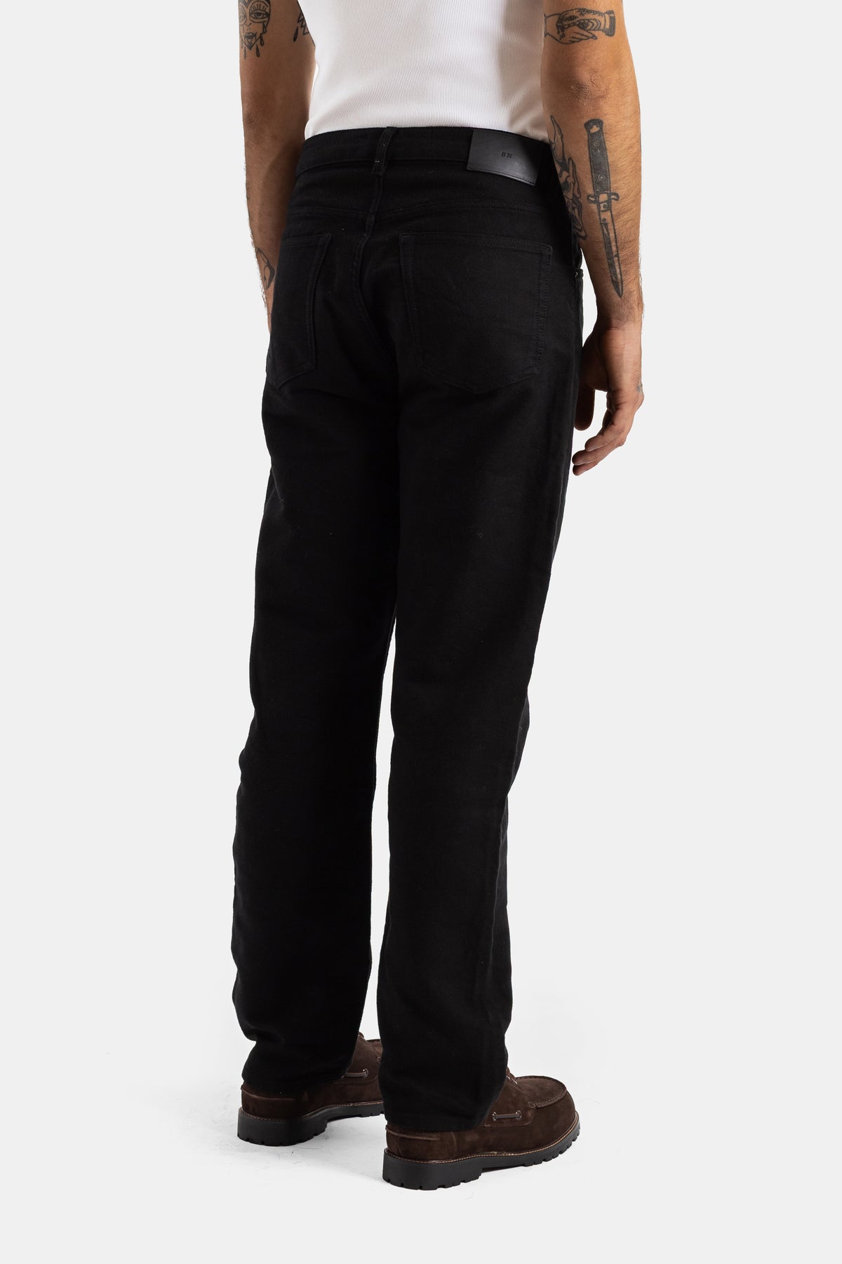 Studio Nicholson men's straight black jeans from the back, featuring leather belt and brown suede soles.