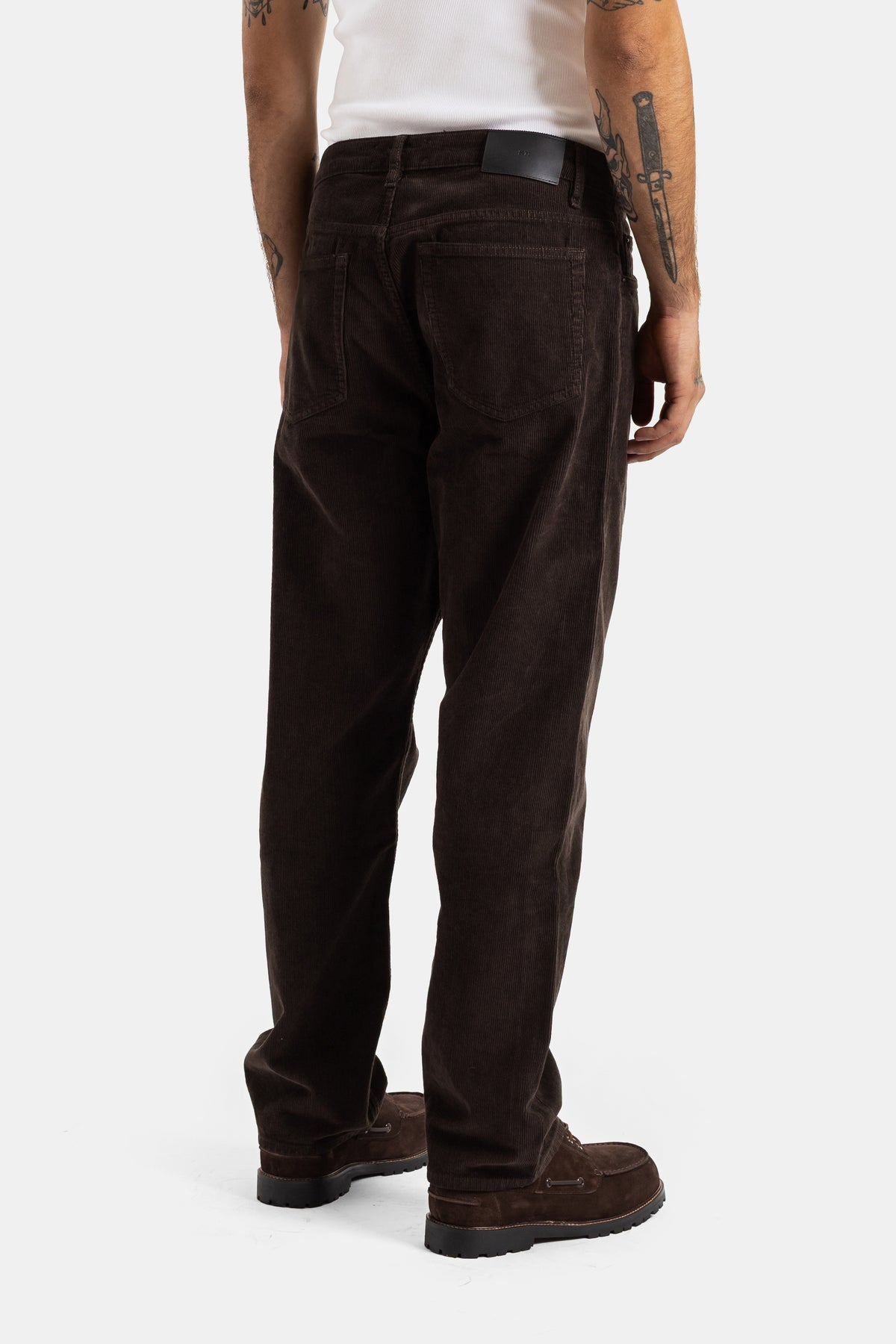 Back view of Studio Nicholson brown corduroy trousers, featuring rear pockets, a black leather patch and belt detail.