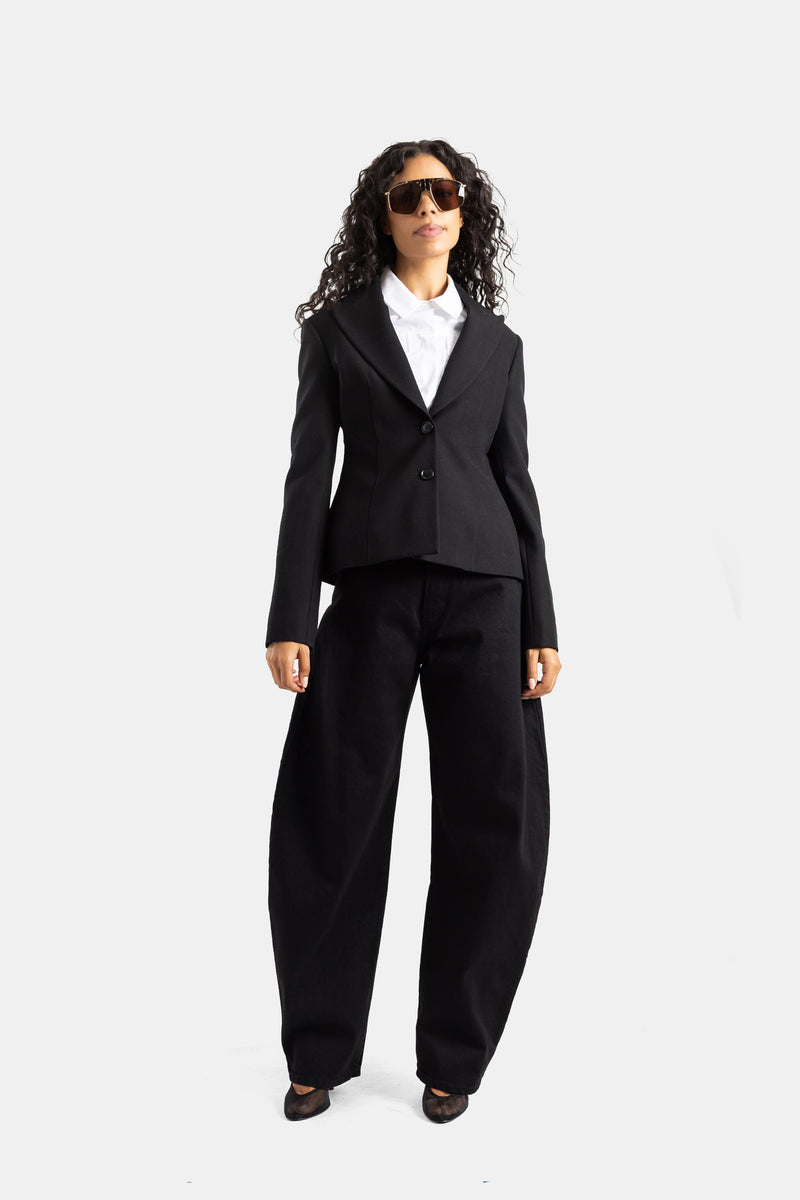 Maison Alaïa（メゾン アライア）の構築的なシルエットの黒いウール製ジャケットに、同色のパラッツォパンツを組み合わせた、シックで洗練されたオールブラックのセットアップを身にまとった女性モデルの全身カット。