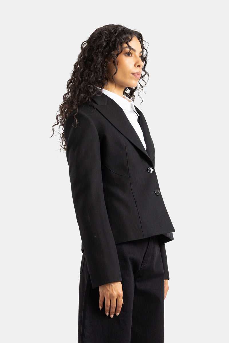 Blazer da donna nero Maison Alaïa indossato di profilo con camicia bianca e pantaloni larghi abbinati