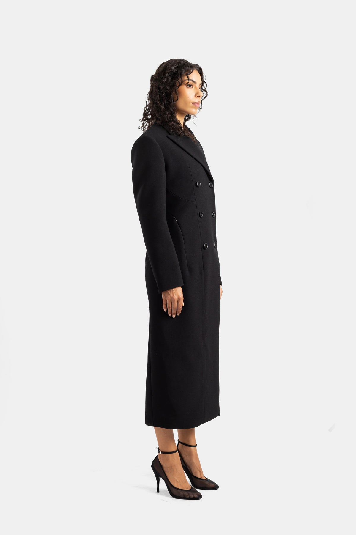 Maison Alaïa slim silhouette black wool coat for women, styled in profile with elegant high-heeled stiletto shoes.
