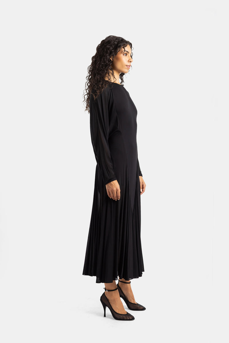 Silhouette di profilo Maison Alaïa: abito nero lungo fluido con maniche lunghe trasparenti e gonna plissettata svasata.