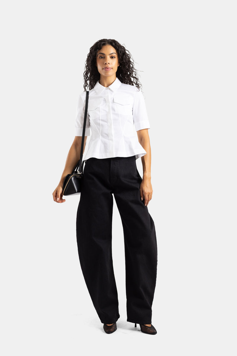 Jeans neri a vita alta Maison Alaïa silhouette arcuata indossati con camicia bianca e borsa a tracolla.