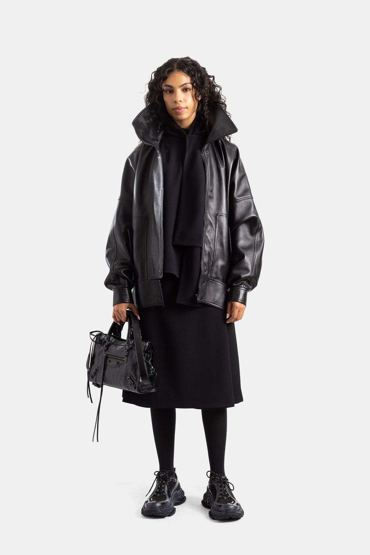 Look total black Balenciaga donna con giubbotto in pelle oversize, borsa nera, sneakers spesse e gonna midi.