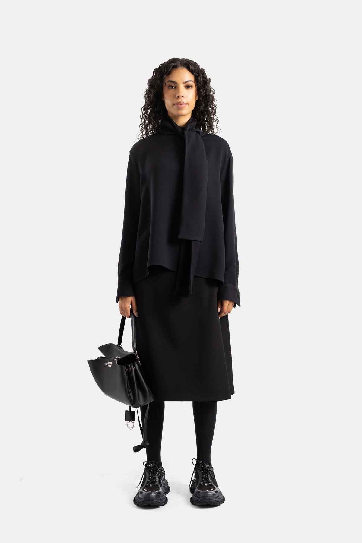Blusa nera Balenciaga con lavallière in un look total black con gonna midi, legging e sneakers chunky.