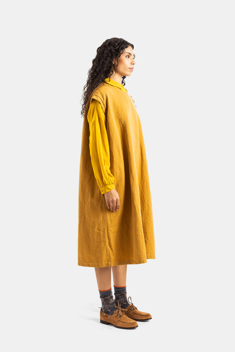 Vista de perfil do vestido Casey Casey Valeme em algodão mostarda com silhueta trapézio sobre camisa amarela.