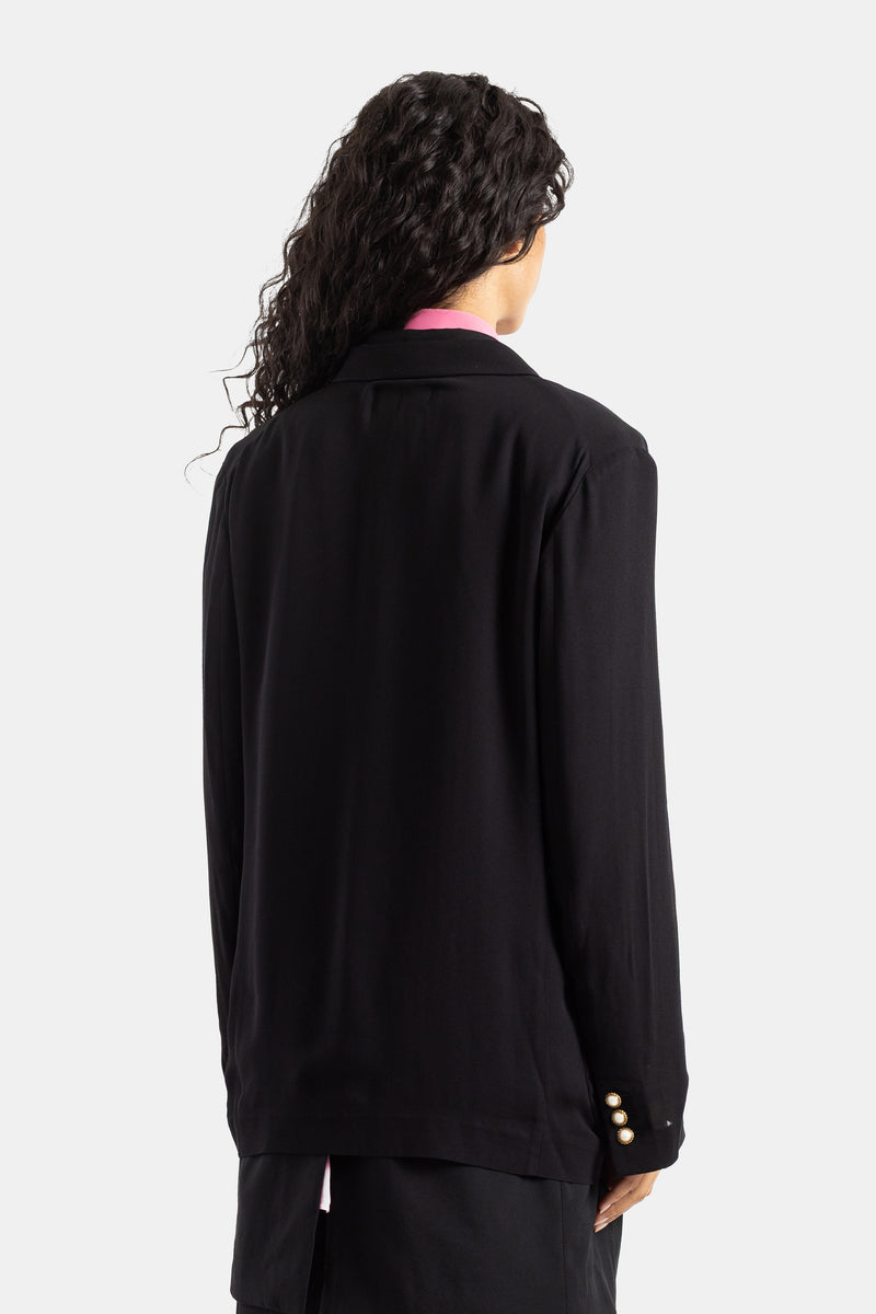 Schwarze, fließende Comme des Garçons Jacke von hinten gesehen, lockiges Haar, Goldknöpfe am Handgelenk.