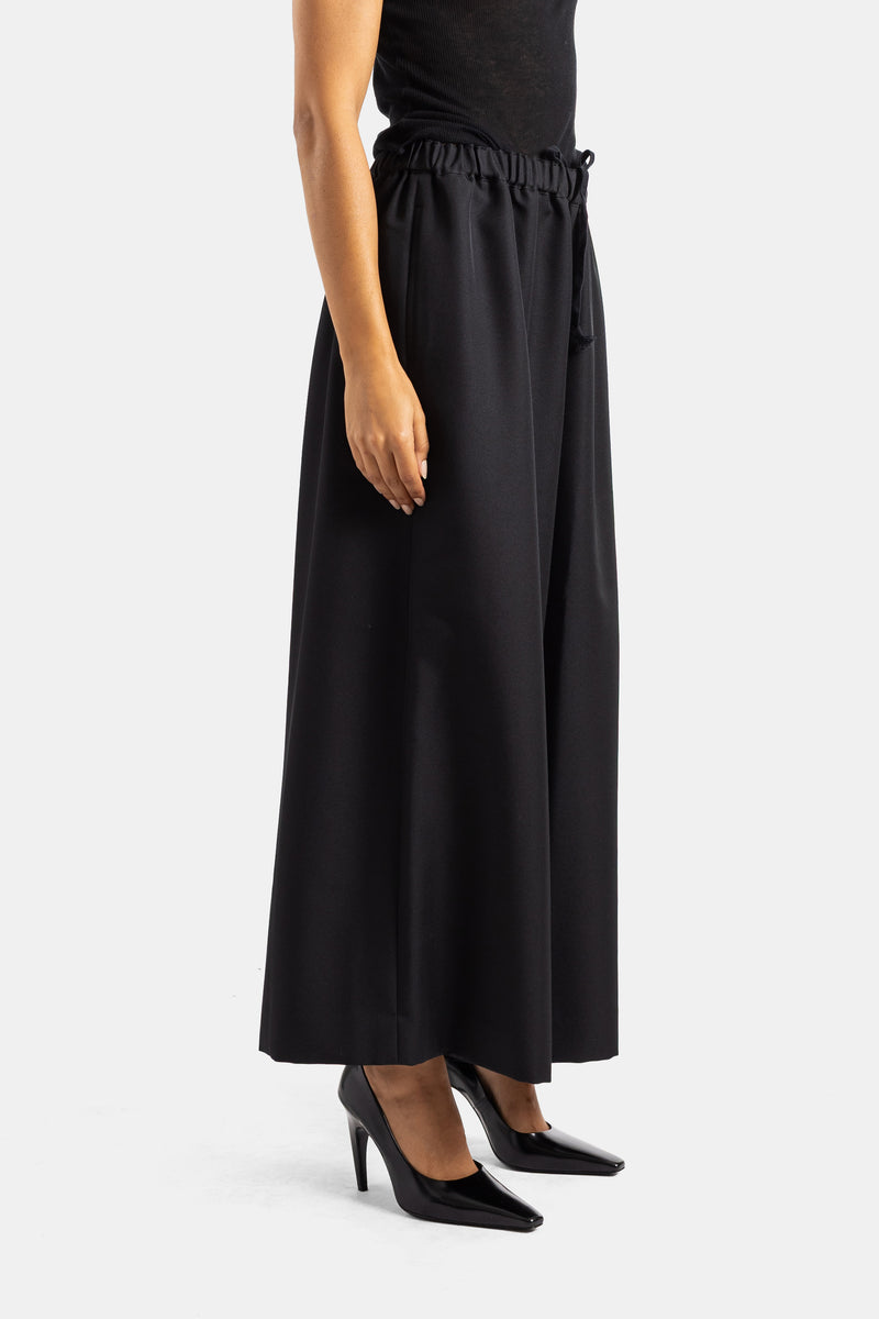 Seitenansicht der schwarzen Comme des Garçons Wide-Leg-Hose mit Raffung am Bund und schwarzen Lack-Pumps.