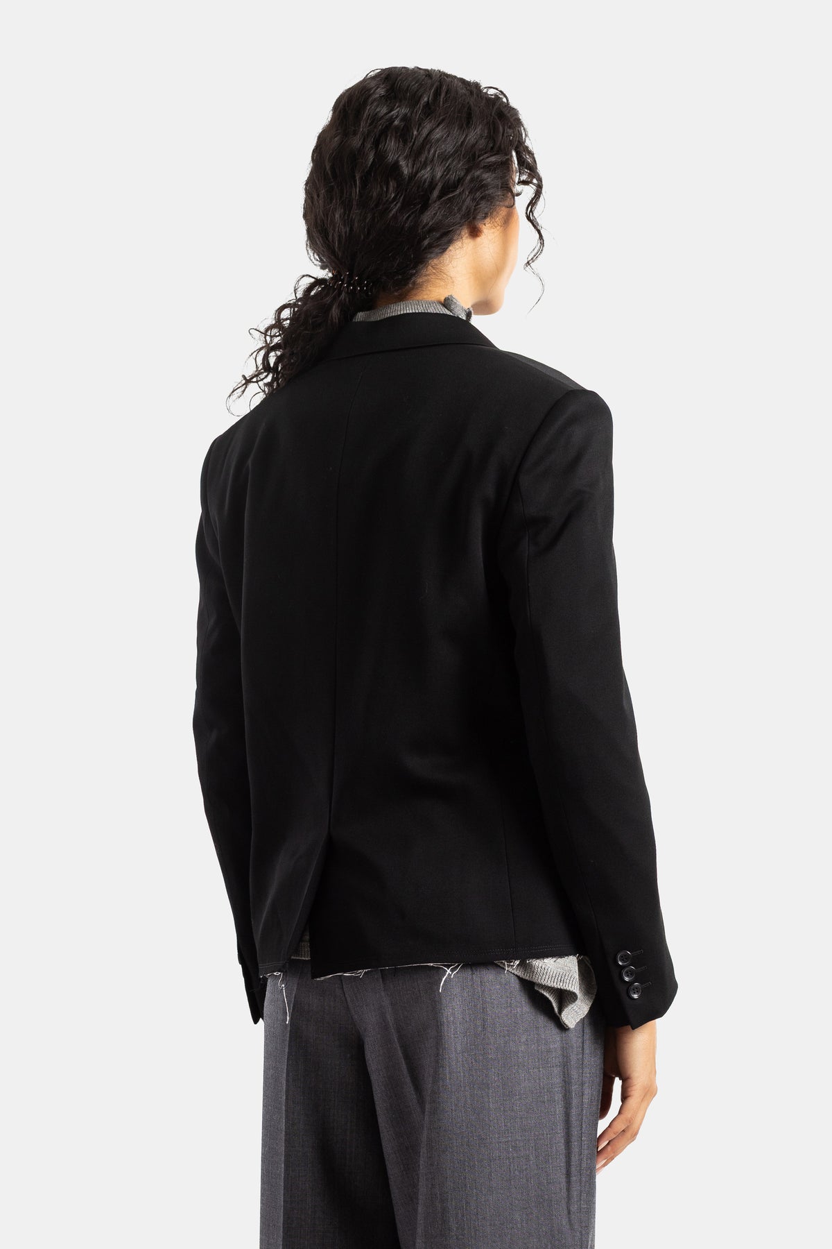 Vista trasera de la chaqueta sastre Comme des Garçons de lana negra con un corte entallado y botones negros.