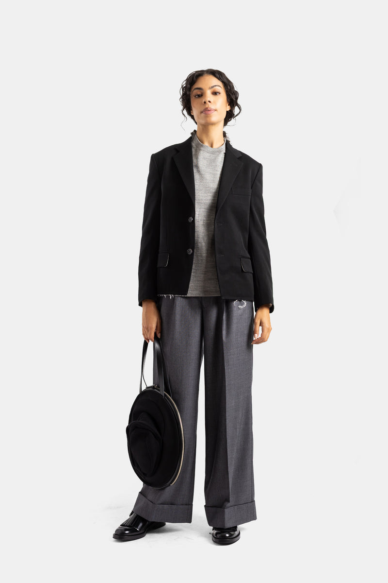 Blazer negro de mujer Comme des Garçons abierto sobre jersey gris, pantalón ancho y bolso redondo negro.
