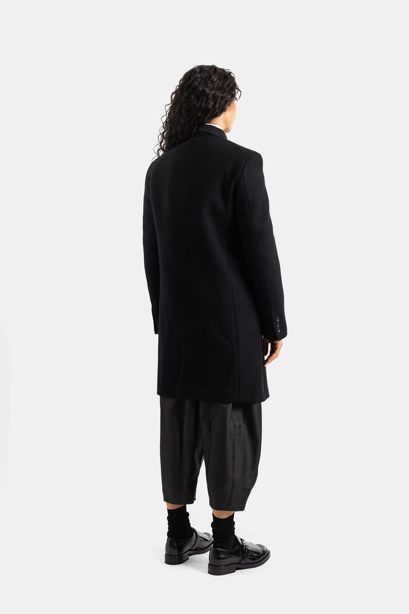 Vista trasera del abrigo de lana negra Comme des Garçons con corte ajustado, zapatos de charol y mocasines.