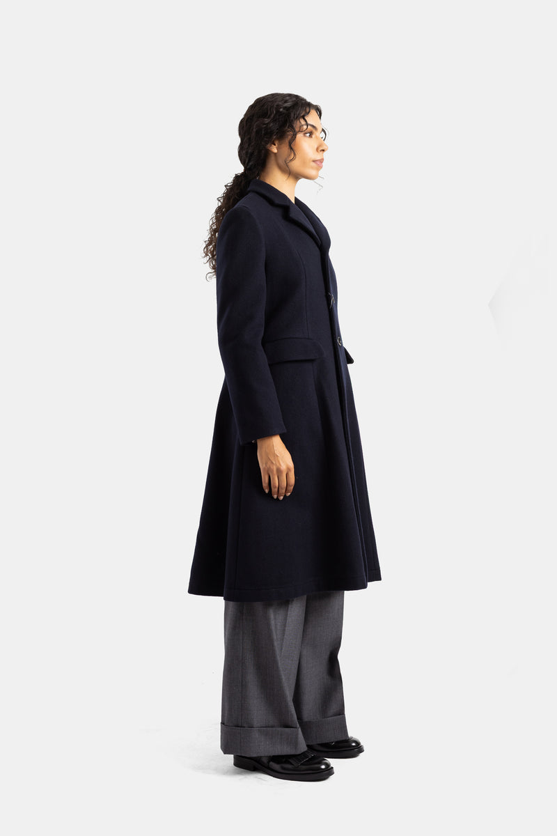 Abrigo de lana azul marino Comme des Garçons de perfil, silueta acampanada a media pierna y bolsillos de parche.