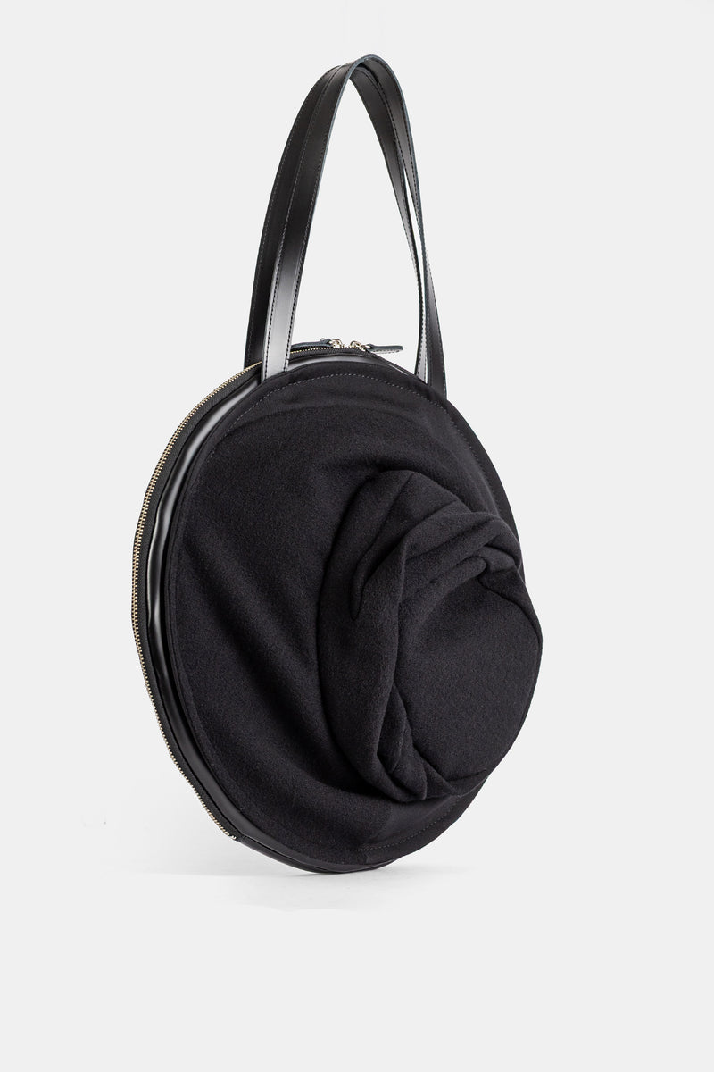 ジュンヤワタナベの円形トートバッグ。ゴールドのファスナーと、黒いフェルトで表現されたハット風の彫刻的なドレープが魅力。洗練されたスタイルとモダンな機能性を融合した、大人のためのシックなラグジュアリーアイテム。