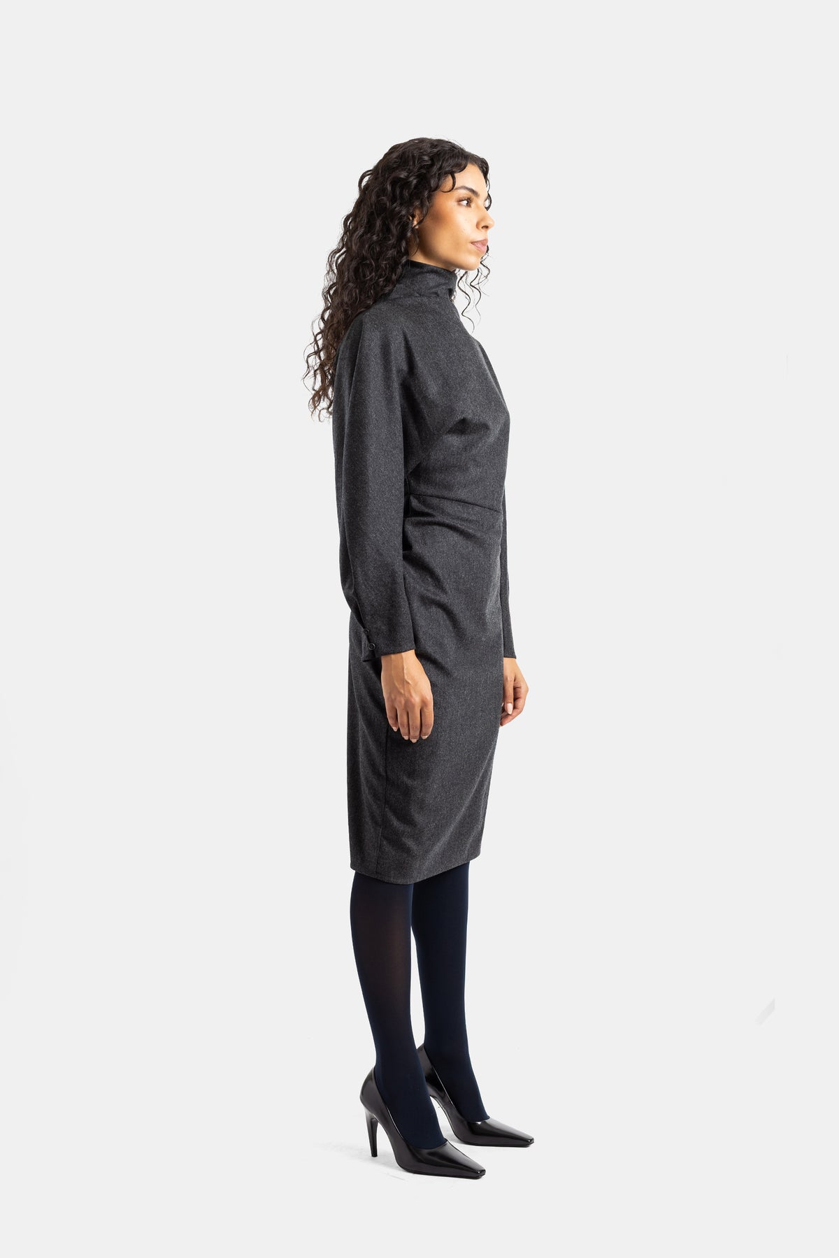 Khaite Harlin-jurk in zijprofiel, midi-silhouet van antracietgrijze wol met zwarte panty en lakleren pumps.