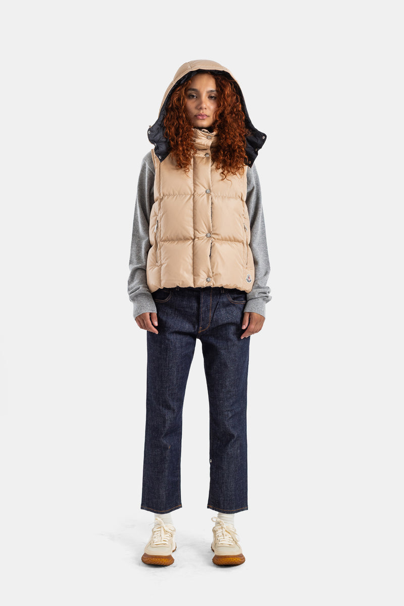 Moncler Macareuxベージュダウンベストを、グレーのセーターとリジッドデニムに合わせた女性のコーディネート。フードを被ることで、ストリート感とハイエンドなラグジュアリーが調和した都会的なルックを演出。