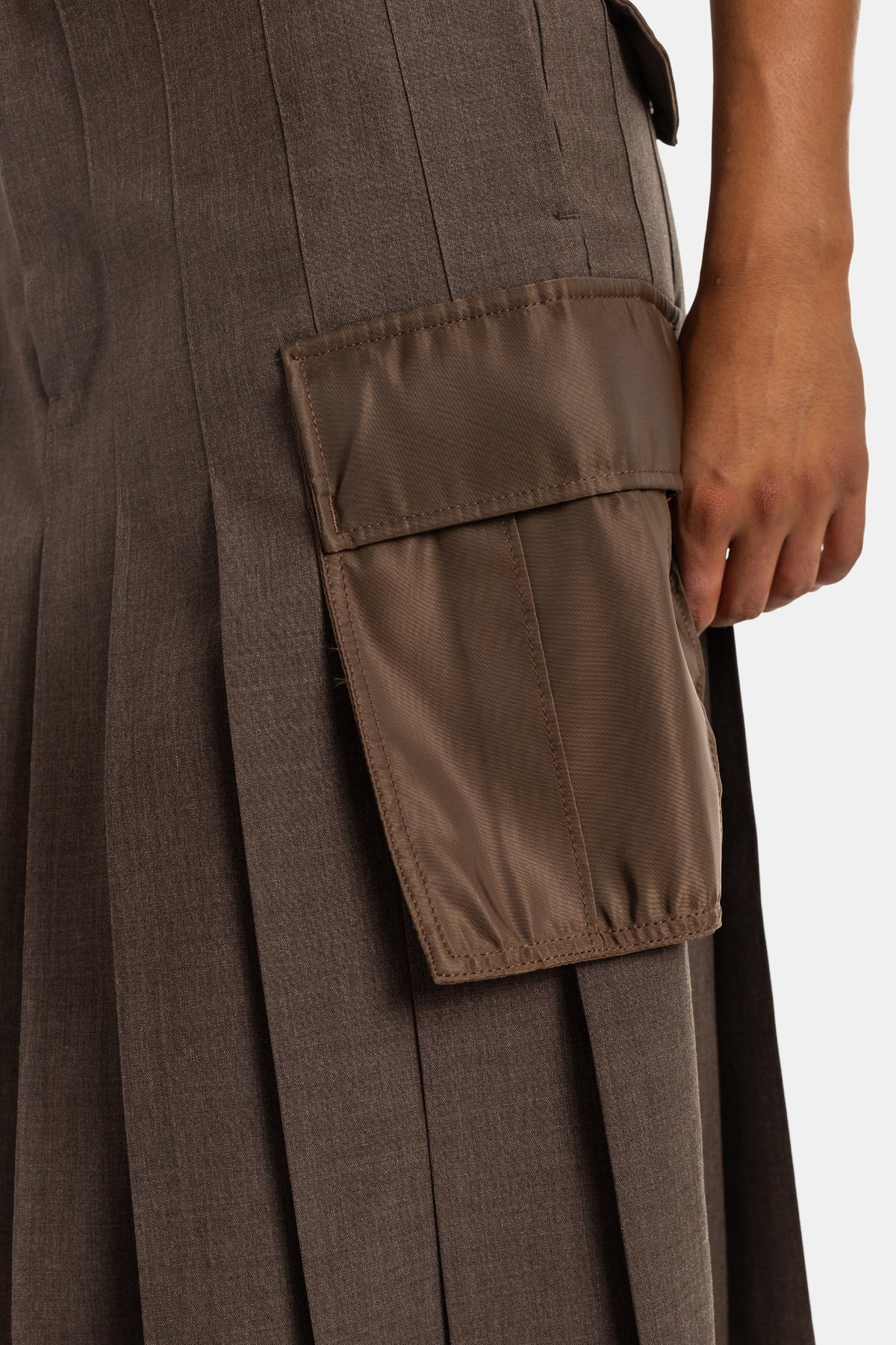 Zoom do bolso cargo aplicado na saia plissada castanha Sacai em nylon acetinado contrastando com o tecido de lã