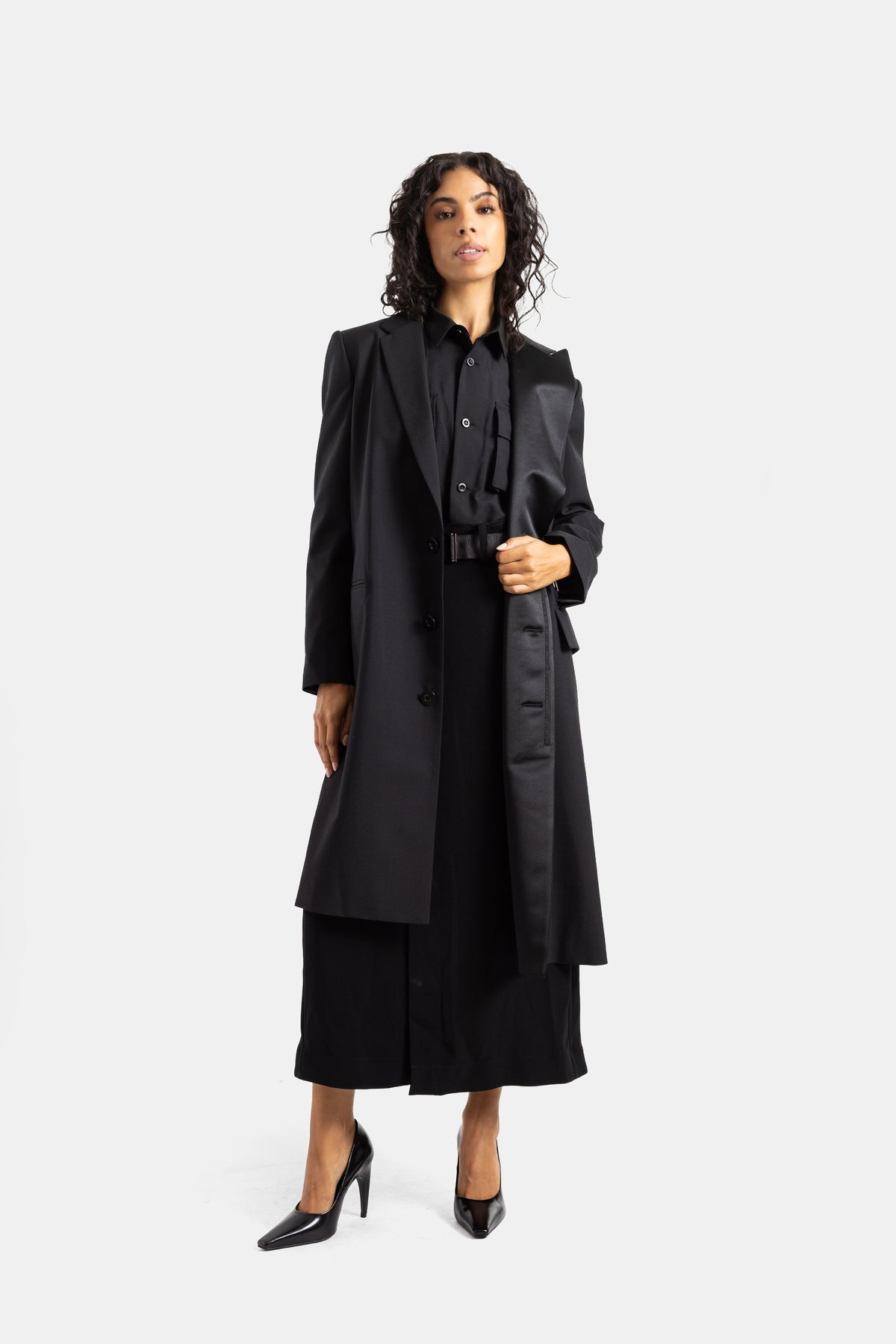 Cappotto nero Sacai frontale con cintura nera con fibbia e pannello asimmetrico in raso cadente sul lato.