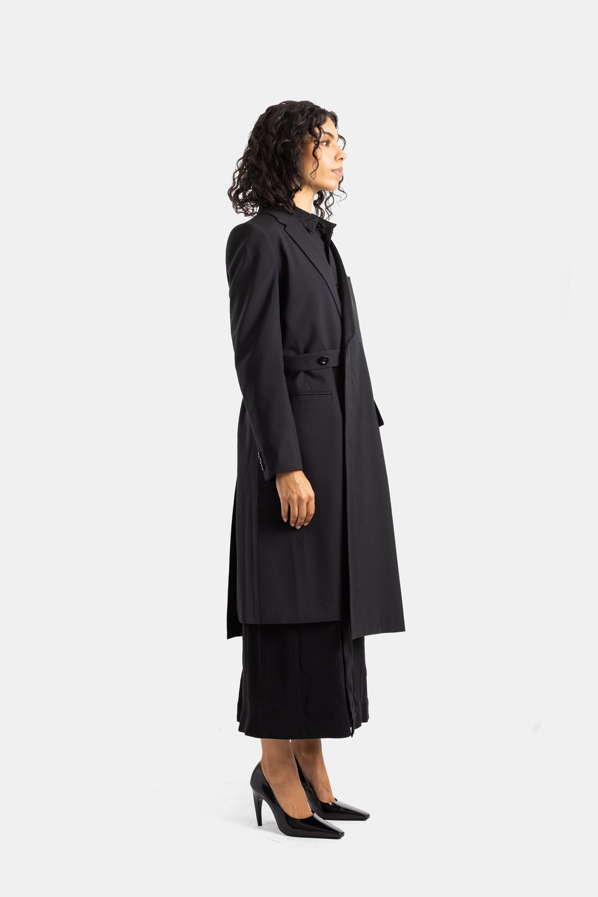 Cappotto nero Sacai di profilo, silhouette lunga e strutturata con bottone singolo e gonna plissettata visibile.