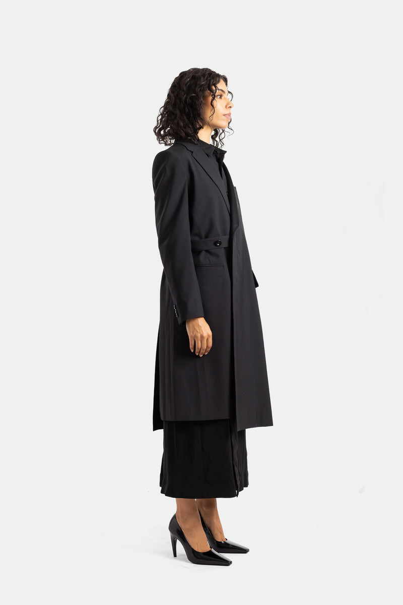 Cappotto nero Sacai di profilo, silhouette lunga e strutturata con bottone singolo e gonna plissettata visibile.