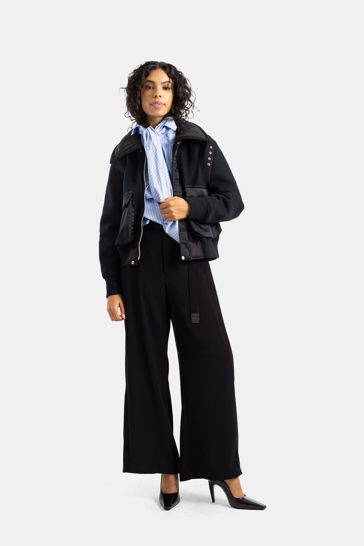Calças largas Sacai pretas com casaco bomber e camisa às riscas azuis, num visual total black estruturado.