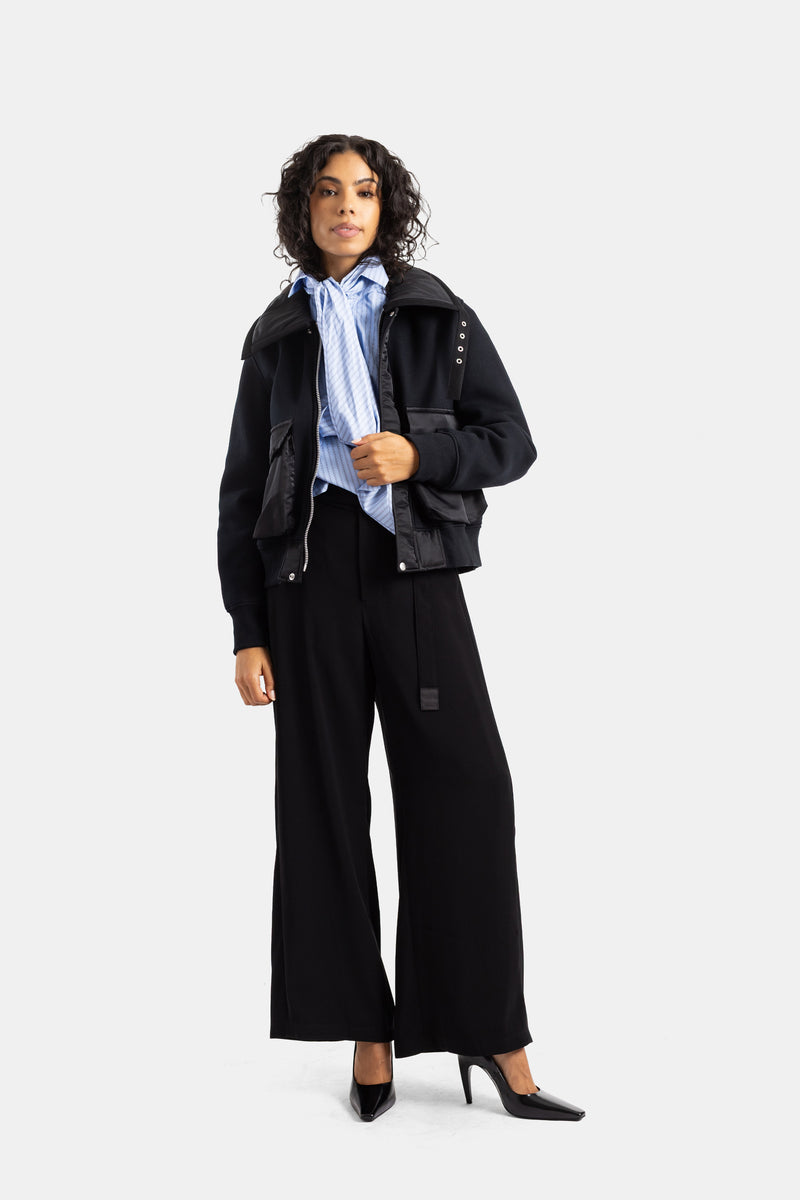 Pantaloni larghi neri Sacai indossati con bomber e camicia a righe blu, per un look total black strutturato
