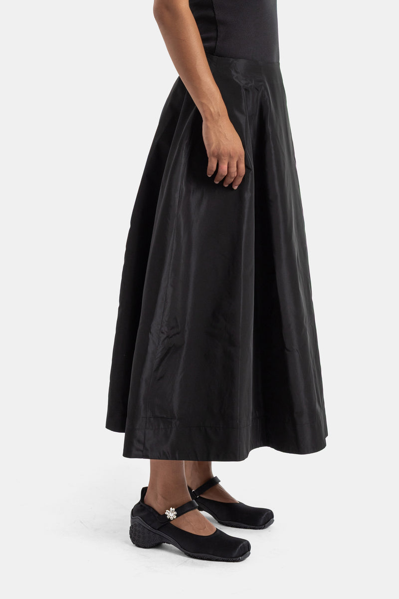 Side view of Simone Rocha black midi skirt in shiny flared taffeta, styled with velvet Mary Jane shoes