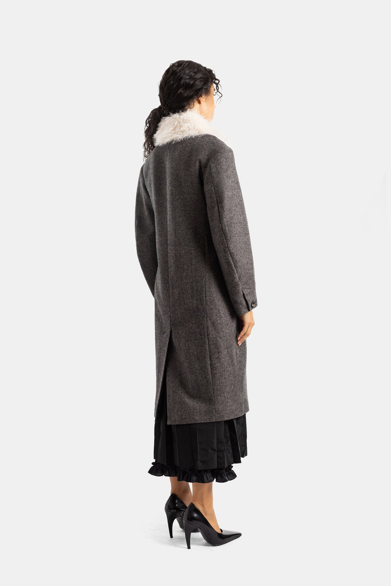 Back view of Simone Rocha's long grey flecked wool coat with ivory fur collar, central vent, black dress and pumps.