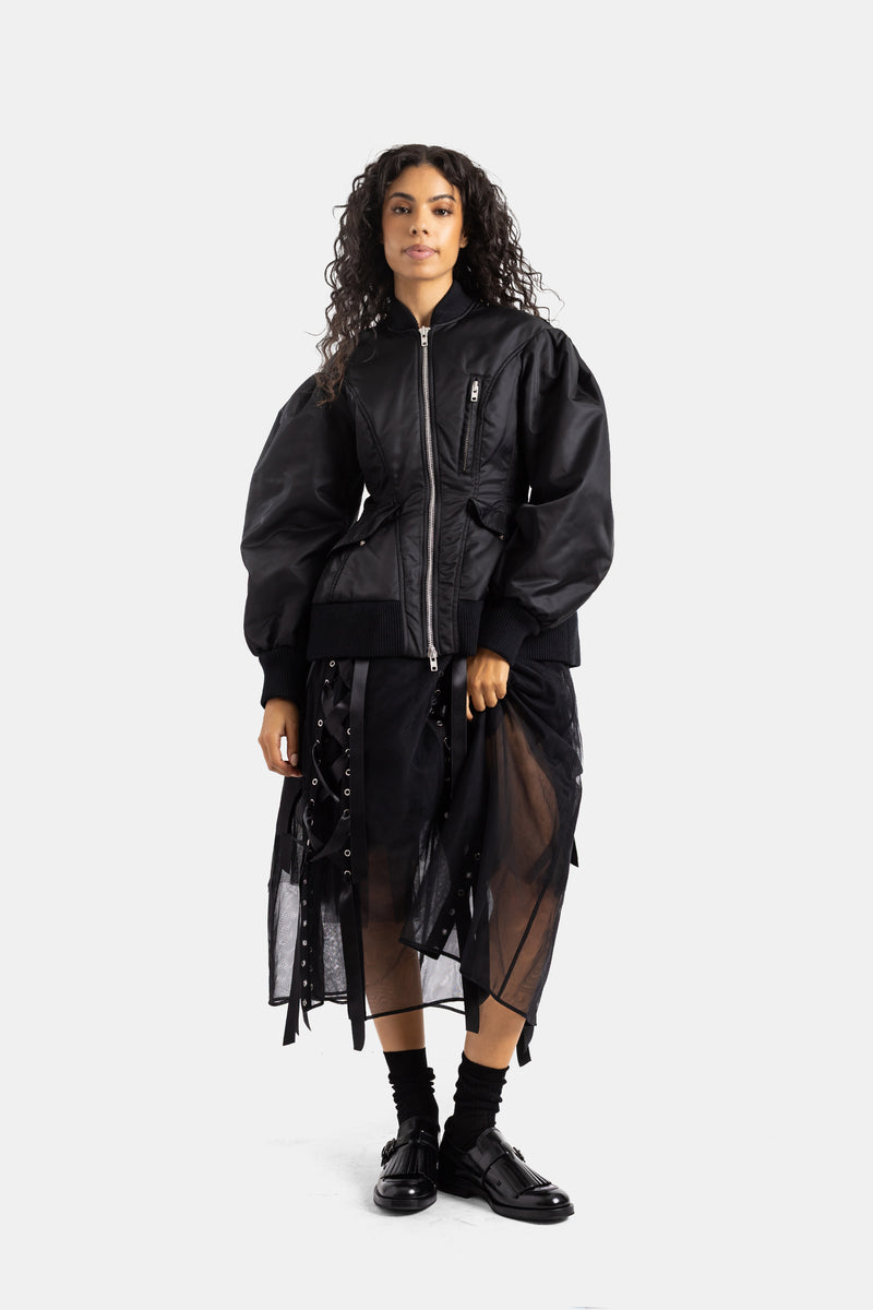 Simone Rocha（シモーネ ロシャ）のブラックボンバージャケットのフロントスタイル。レースアップコルセットに、透け感のあるオーガンジースカートとモンクシューズを合わせた、モードな女性向けコーディネート。