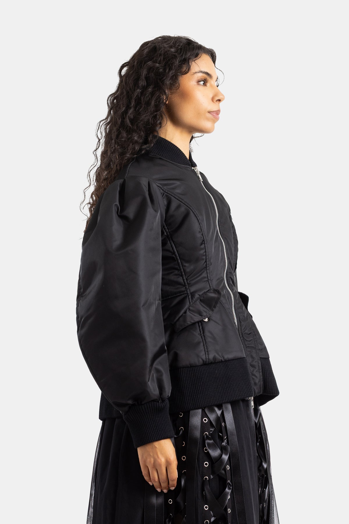 Simone Rocha（シモーネ ロシャ）の黒のボンバージャケットを着用した女性のサイドプロフィール。シルバーのフロントファスナーが輝き、チュールスカートをレイヤードした洗練されたエディトリアルルック。
