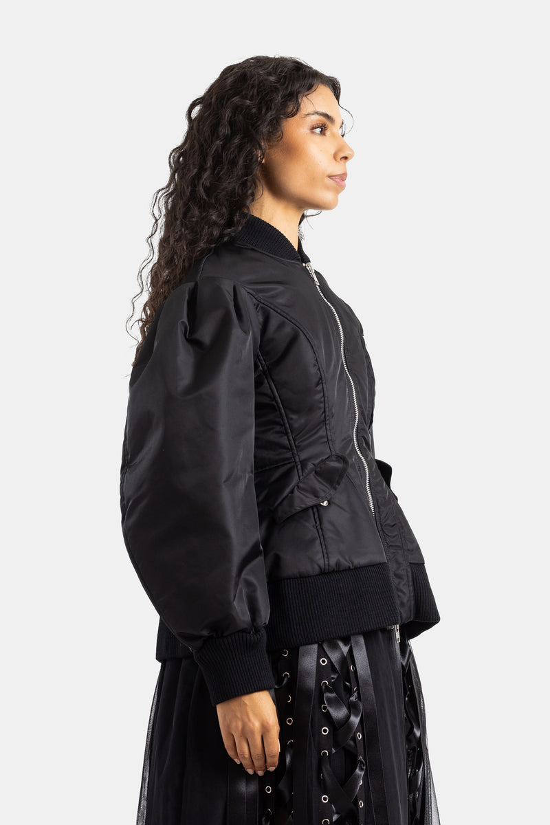 Simone Rocha（シモーネ ロシャ）の黒のボンバージャケットを着用した女性のサイドプロフィール。シルバーのフロントファスナーが輝き、チュールスカートをレイヤードした洗練されたエディトリアルルック。